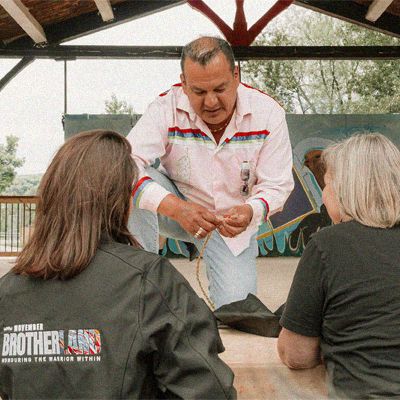 Photo of a man demonstrating traditional Indigenous crafts to two seated people.